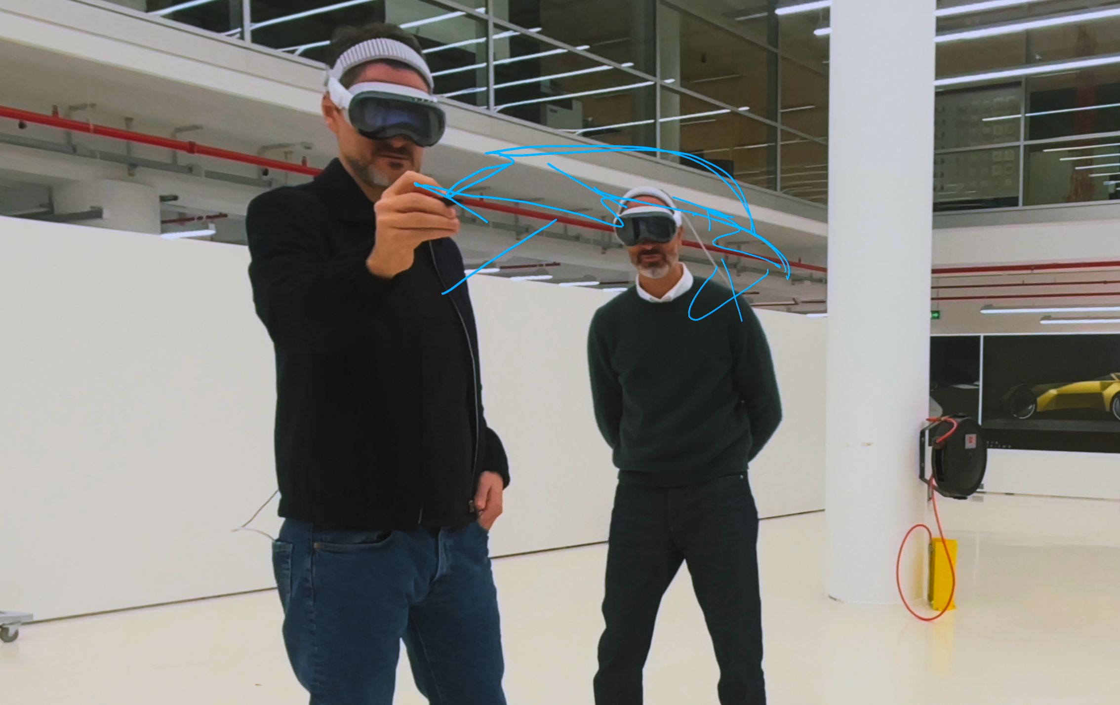 Two designers wearing Apple Vision Pro in a studio, one sketching a free-hand blue spline annotation in mid-air with a stylus.